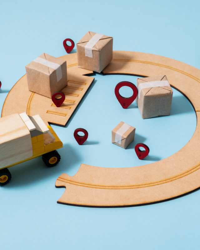 Construction Supply Chain Southeast Asia symbolises by wooden toy truck on circular road with packages and location pins on blue background.