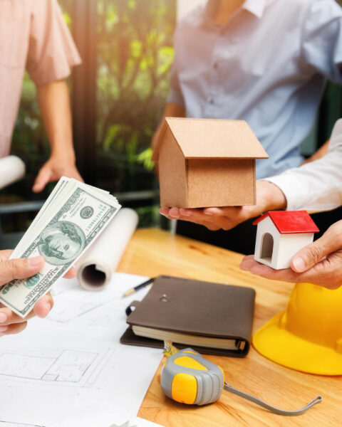 Individuals exchanging money for a small house model with architectural plans and helmets on the table, symbolising Southeast Asia Construction Project Financing.