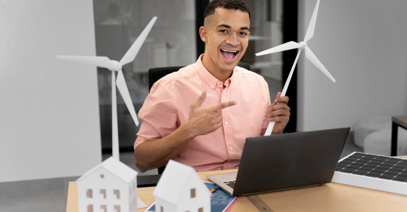 Renewable Energy in Southeast Asia Construction represented by Person holding a wind turbine model at a desk with laptop and miniature houses.